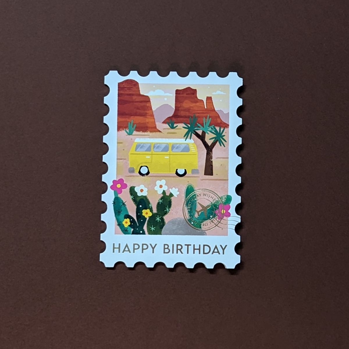 Carte d'anniversaire ornée d'un camping-car de style vintage dans le parc national de Joshua Tree, ce design élégant est découpé à l'emporte-pièce en forme de timbre et orné d'un cachet de la poste doré et de l’inscription Happy Birthday..