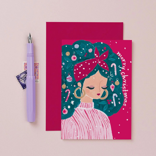 Carte Merry Christmas avec l’illustration d'une jeune fille dont les cheveux sont joliment décorés de sucres d’orge, de boules de Noël et de houx créant un look festif et amusant .