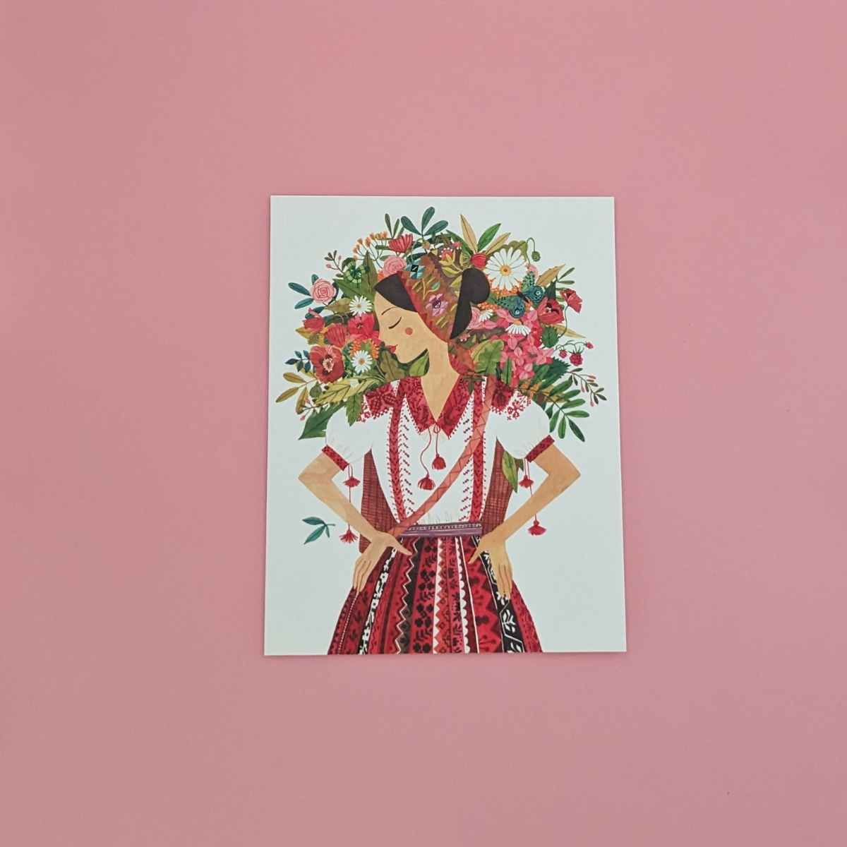 Une jeune roumaine vêtue d'une tenue traditionnelle, porte à son dos un panier rempli de jolies fleurs. Cette carte évoque la beauté, la nature et la tradition.