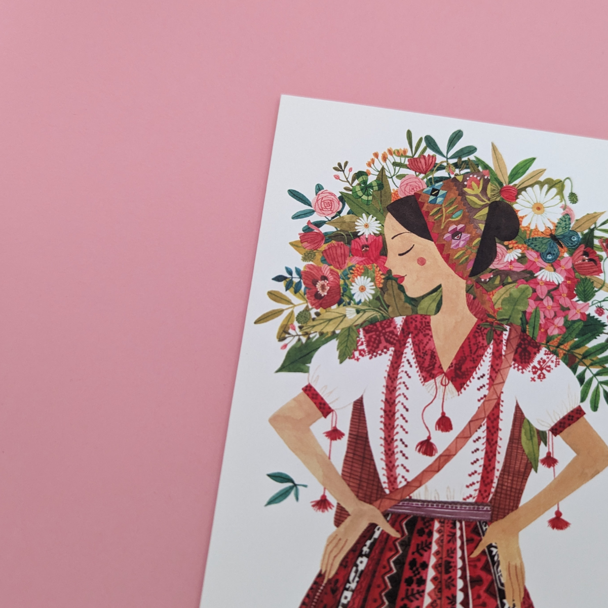 Une jeune roumaine vêtue d'une tenue traditionnelle, porte à son dos un panier rempli de jolies fleurs. Cette carte évoque la beauté, la nature et la tradition.