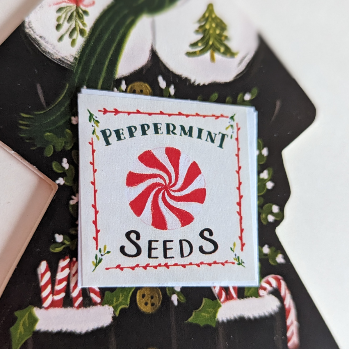 Carte de voeux découpée en forme de personne. Représentation d'une femme qui porte un manteau d'hiver avec des sucres d'orge qui sortent de ses poches. Un petit sachet de graines de menthe poivrée est collé sur le devant de la carte comme si ce petit personnage vous l'offrait.