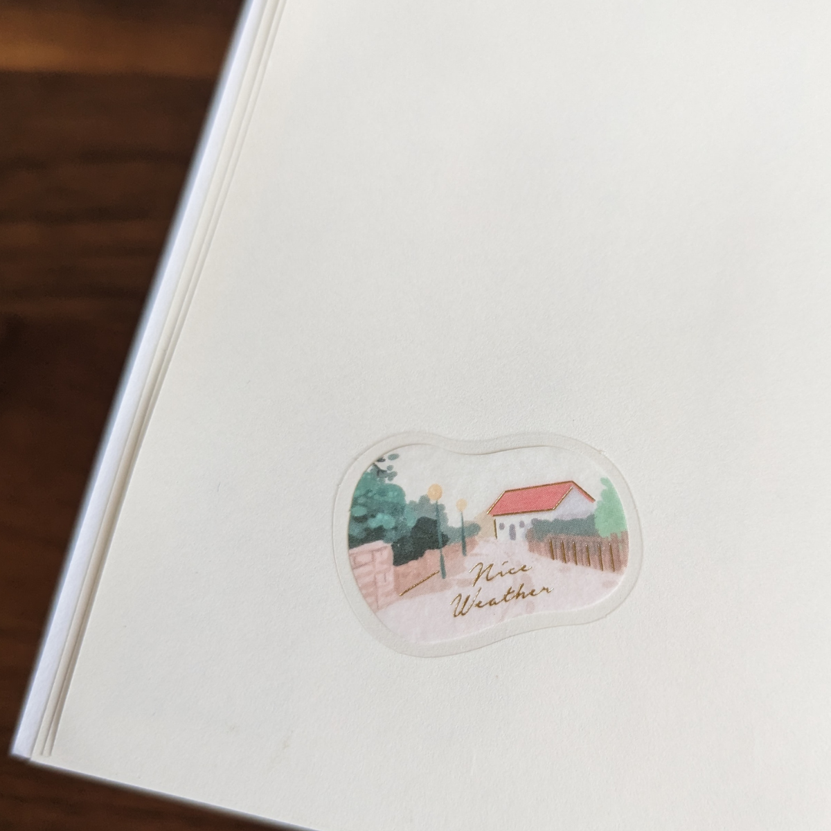 Sticker d'une maison dans une ruelle avec une inscription à l'or chaud : " Nice Weather ".