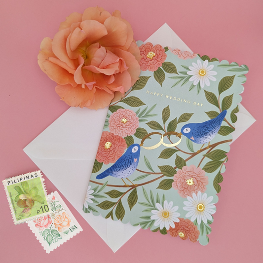 Une illustration poétique de deux oiseaux bleus perchés sur une branche qui s'échangent avec leurs becs des alliances en or, symbole du lien sacré du mariage. Carte découpée à l’emporte-pièce.