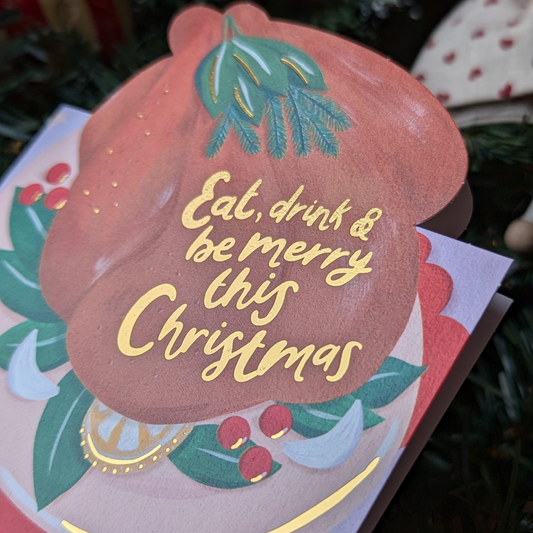 Carte de Noël avec le dessin d'une dinde sur une table et une inscription à l'or chaud " eat, drink and be merry this christmas ".