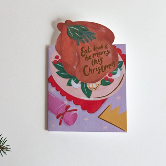 Carte de Noël avec le dessin d'une dinde sur une table et une inscription à l'or chaud " eat, drink and be merry this christmas ".