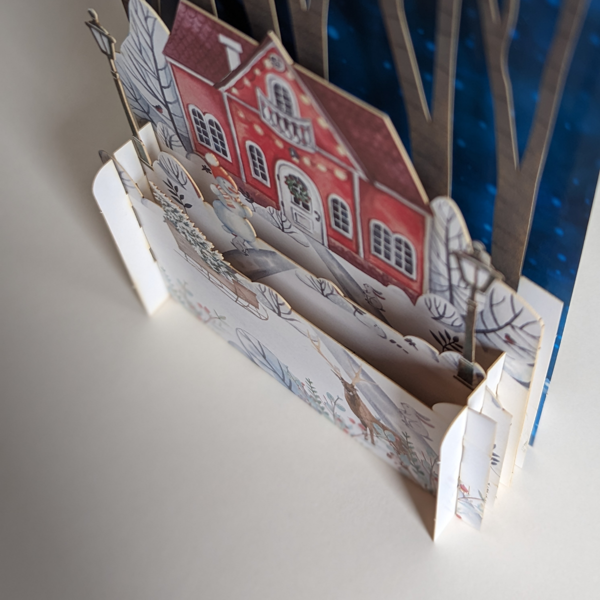 Cette carte en 3D pop-up présente une scène hivernale avec une charmante maison nichée au cœur d'une forêt enneigée. 