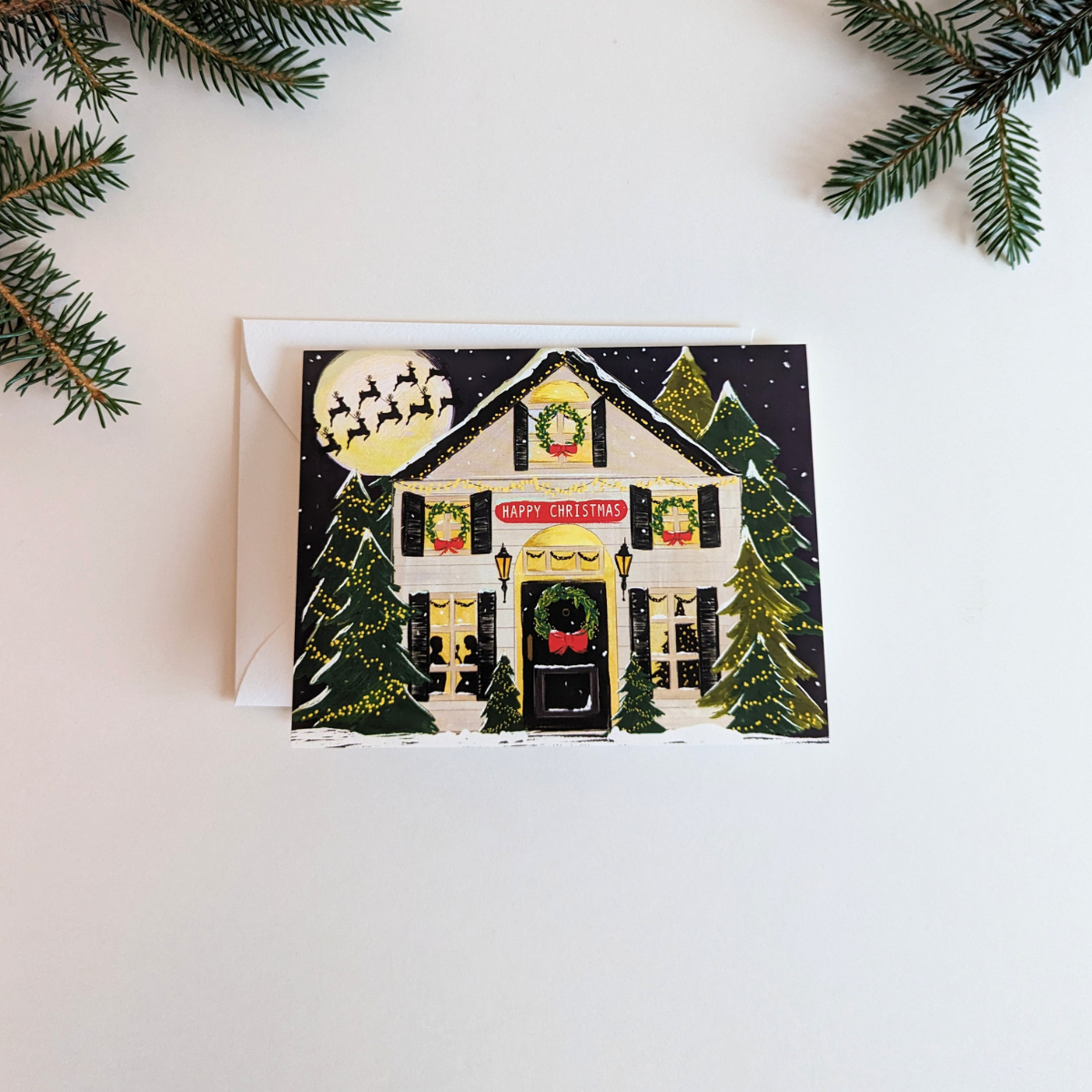 Carte de Noël dessinée par The First Snow. Une jolie maison decorée pour les fêtes, une couronne sur la porte, des guirlandes dans les sapins et les rennes du père-noël dans le ciel.