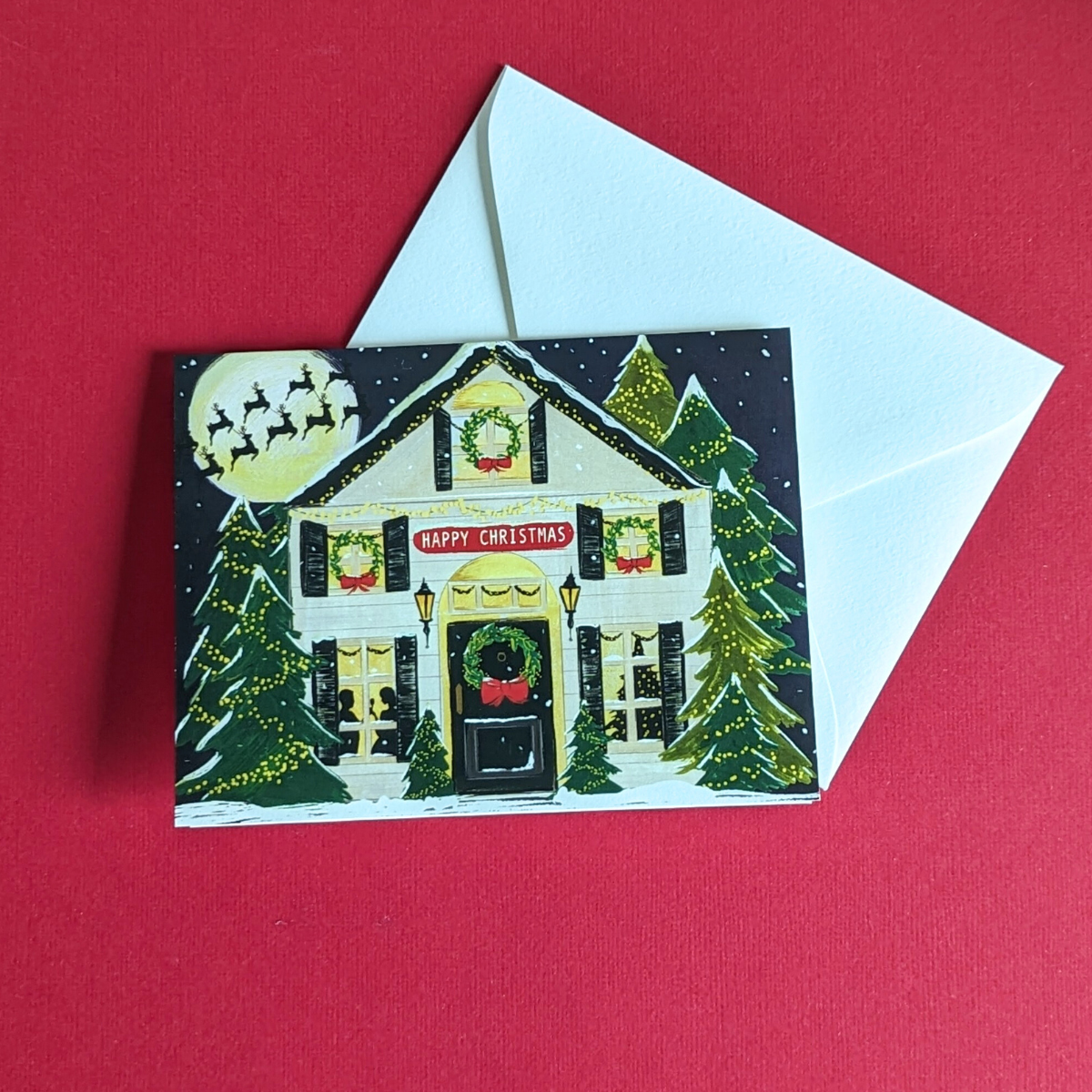 Carte de Noël dessinée par The First Snow. Une jolie maison decorée pour les fêtes, une couronne sur la porte, des guirlandes dans les sapins et les rennes du père-noël dans le ciel.